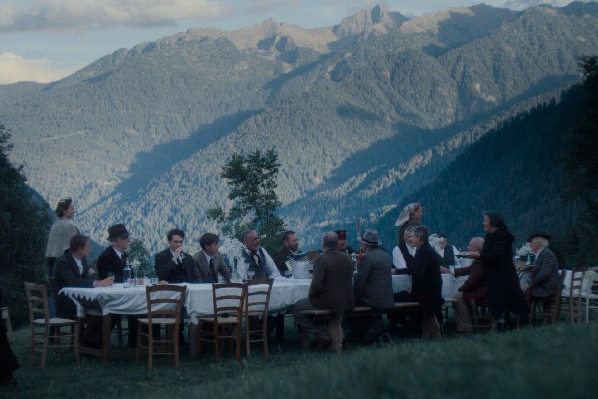 A long table outdoors with people seated and empty chairs against backdrop of wooded mountains.