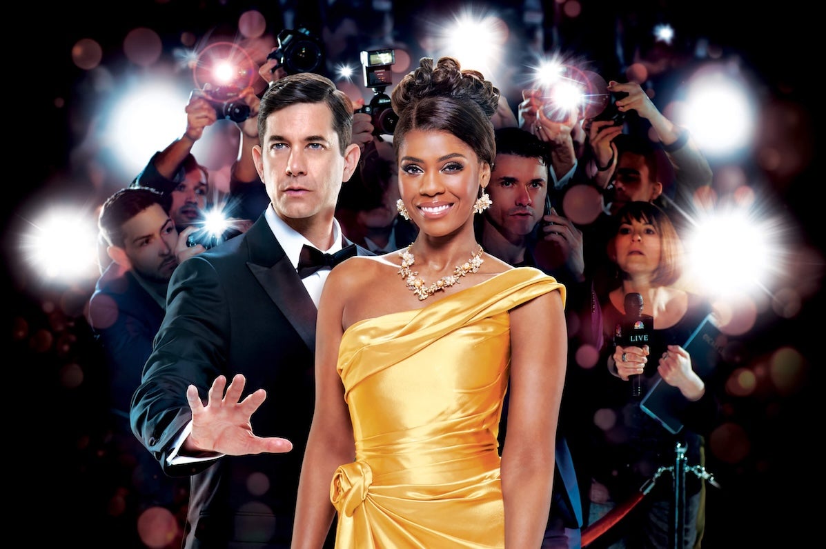 A smiling woman in bright yellow evening gown beside man in tuxedo holding out an open hand in a warning manner