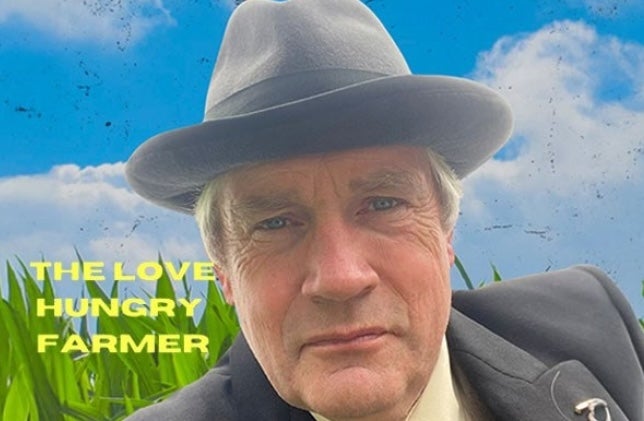 A man in grey brimmed hat and suit and tie with grass behind him.