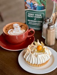 Cappuccino in a brown mug beside a cream tartlet on a white plate