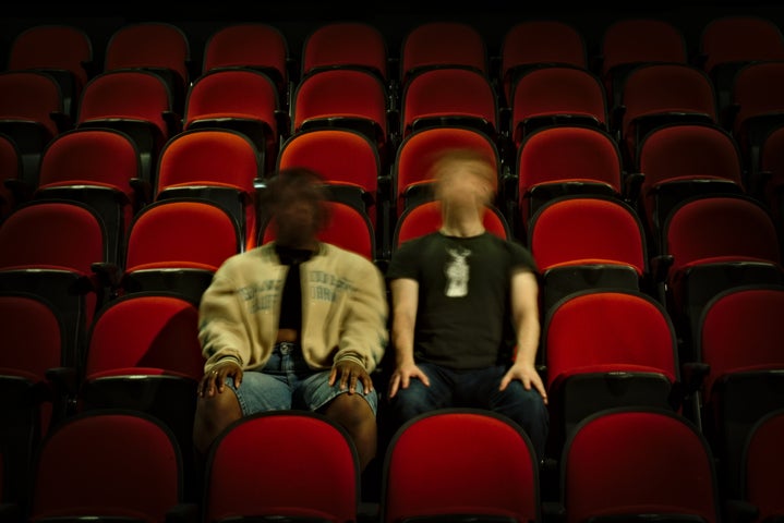 Two people sit on red chairs, their faces are blurry due to motion. They both have their arms stretched out, hands on knees.