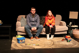 On a stage, 2 people are seated on a sofa with children's toys on a mat in front of them.