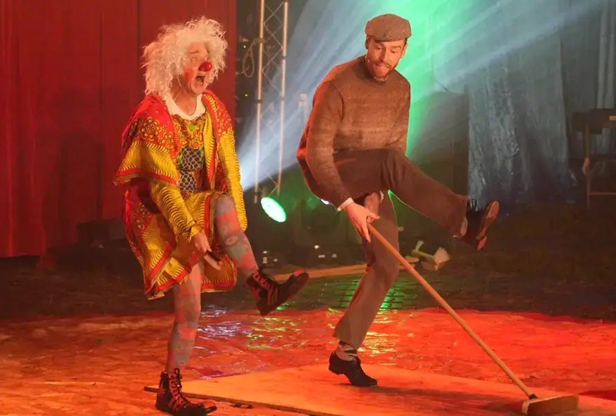 2 people dancing on a stage, one dressed in patterned clothes wearing a white wig and red nose.