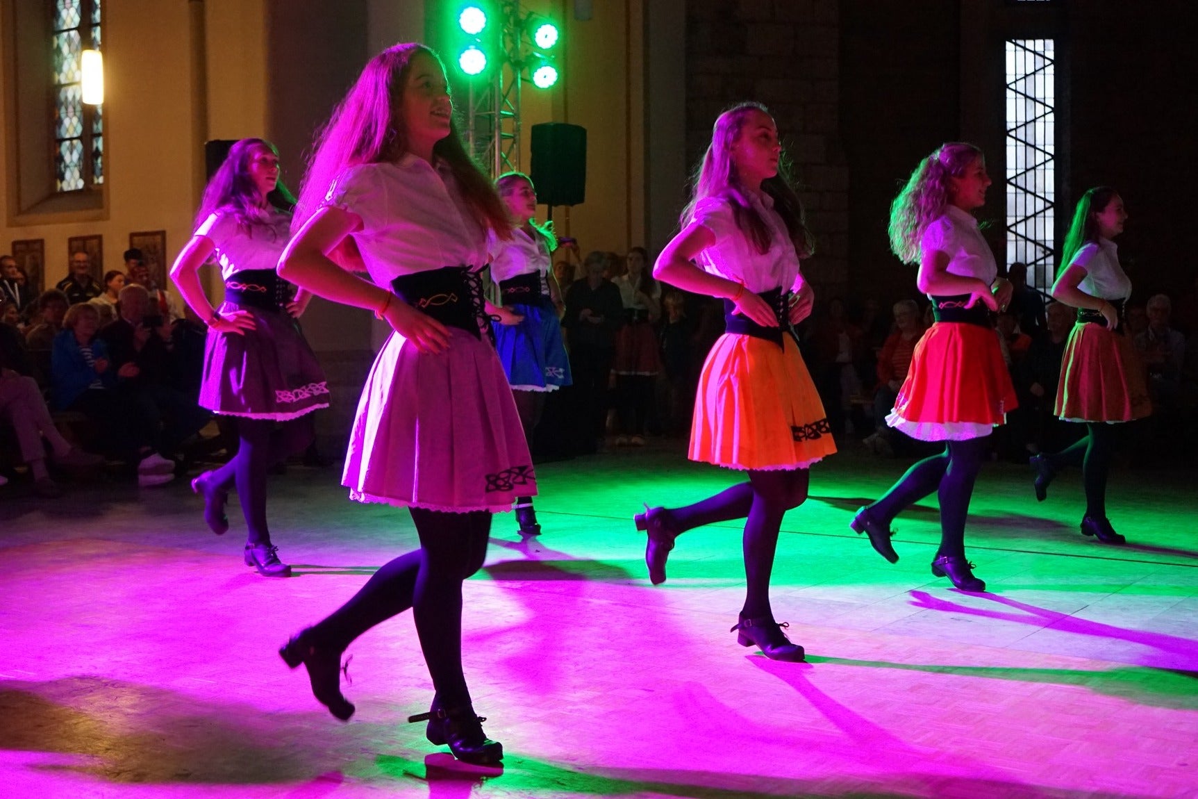 Group of 6 irish dancers spread out in 2 lines in hard shows with audience looking on.