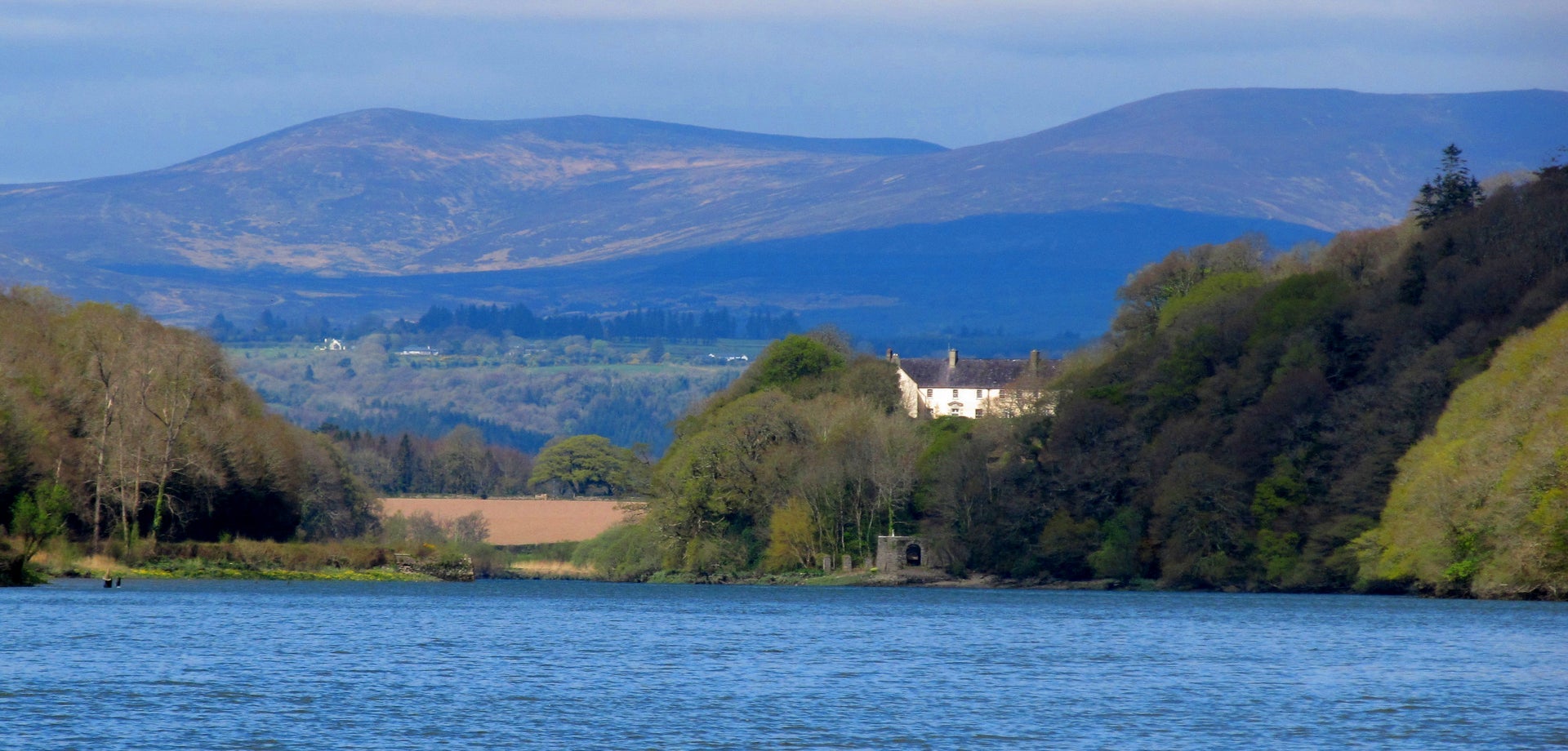 A large period house in the distance surrounded by trees overlooking the sea