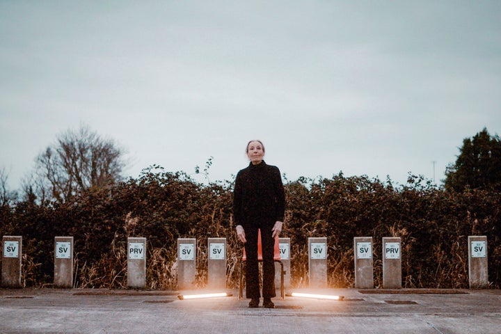 An older woman dressed in black is standing outdoors on a dull day on concrete area with 2 lit fluorescent tubes on the ground either side of her 