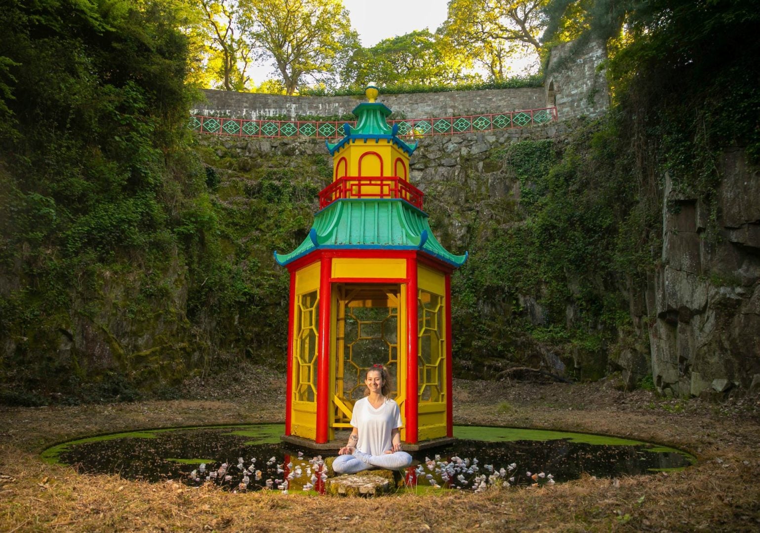 A small yellow and red round pagoda shaped building with green roof surrounded by tall, green covered walls with a person seated in front in a yoga position.