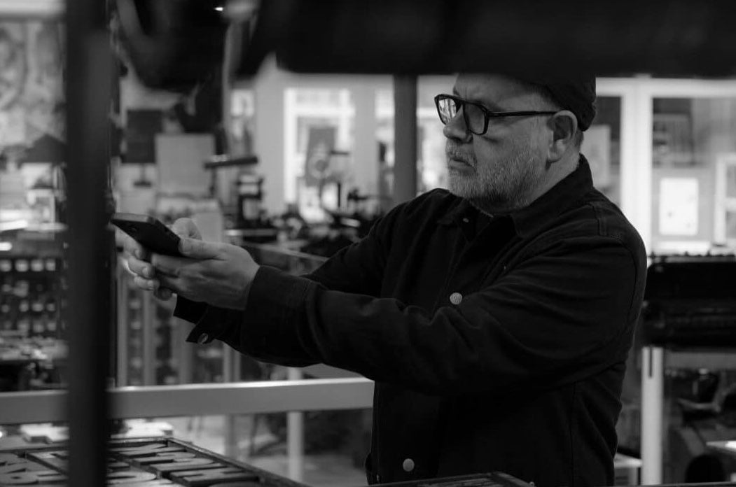 Anthony Burrill and Maser In Conversation, a man in a museum holding his mobile phone horizontally out in front of him with various print machines in the background.