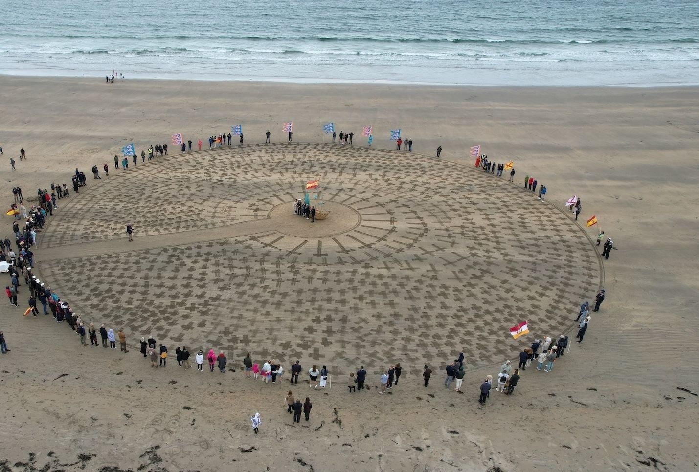 People form a circle around crosses marked in the sand at a beach memorial event