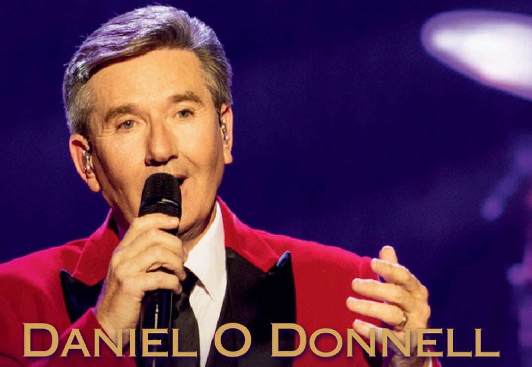 A man in red jacket and black tie is singing into hand held mic against dark purple background.