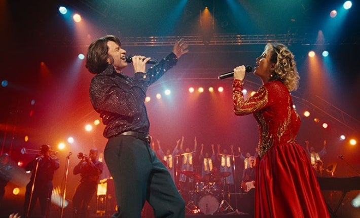 A man and woman singing with passion into mics, facing each other on a stage.