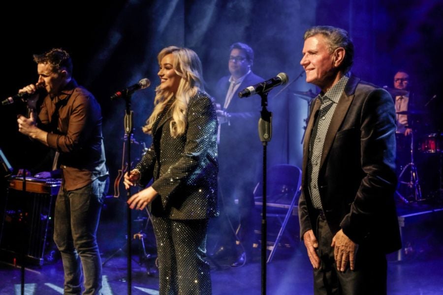 3 people in a line on a stage each singing into a mic on a stand with musicians behind.