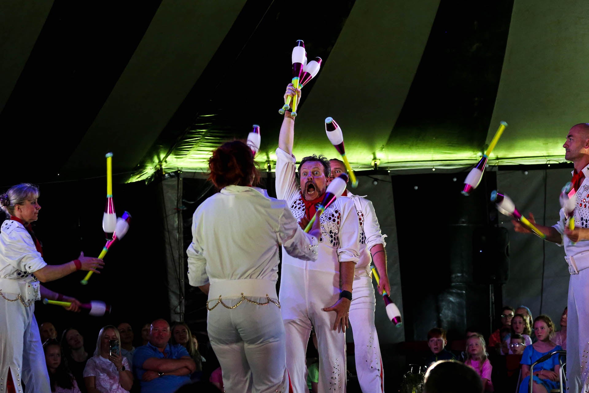 In a dark tent, people dressed in white are juggling in formation with large batons.
