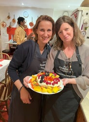 Two ladies in aprons posing with a cake with lots of fruit as decorations