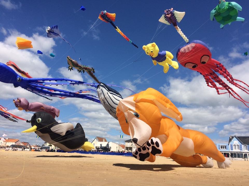 On a sunny day, large kites in the shape of various animals are inflated and hovering just above a large, sandy beach.