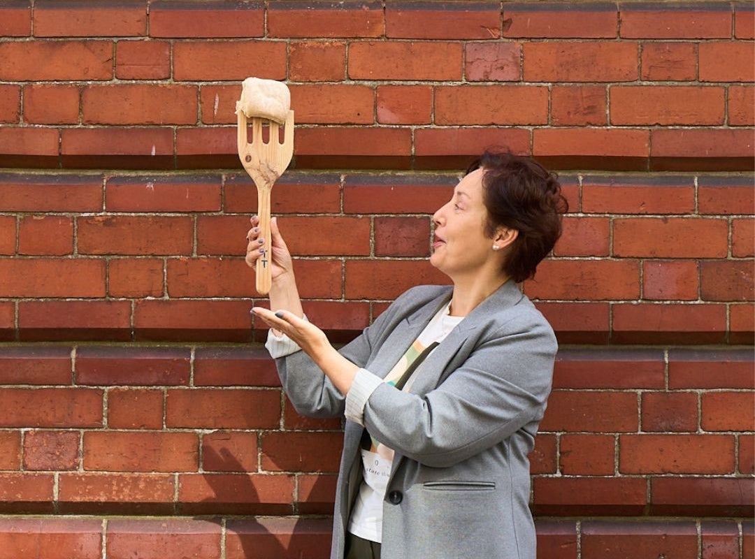 Person in a grey blazer holding a wooden fork in front of a red brick wall