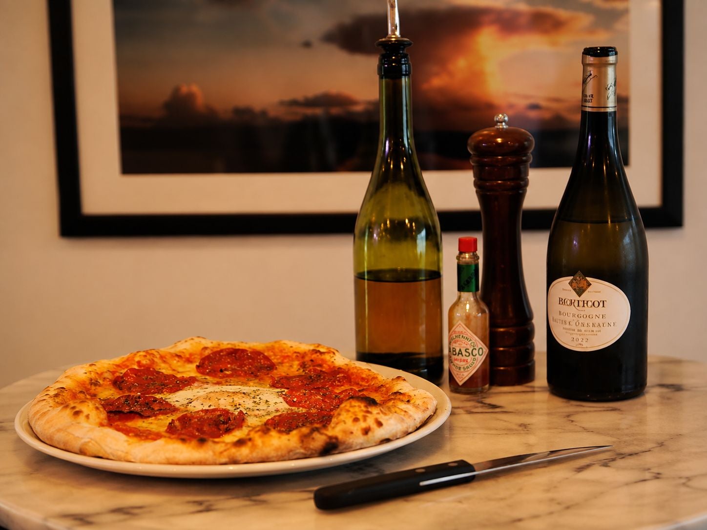 A pizza served alongside a bottle of wine with olive oil and hot sauce