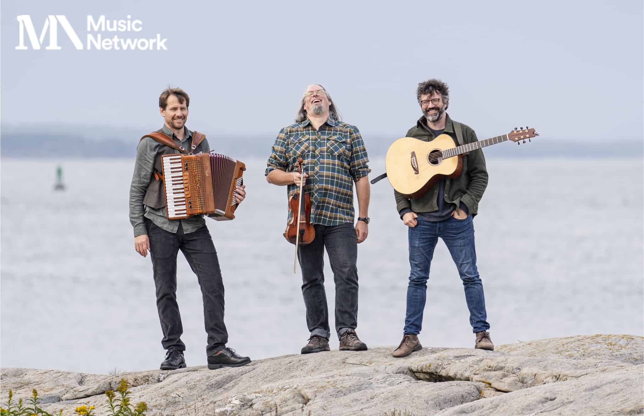 3 smiling men are standing on a large rock with musical instruments against a flat, grey sea.