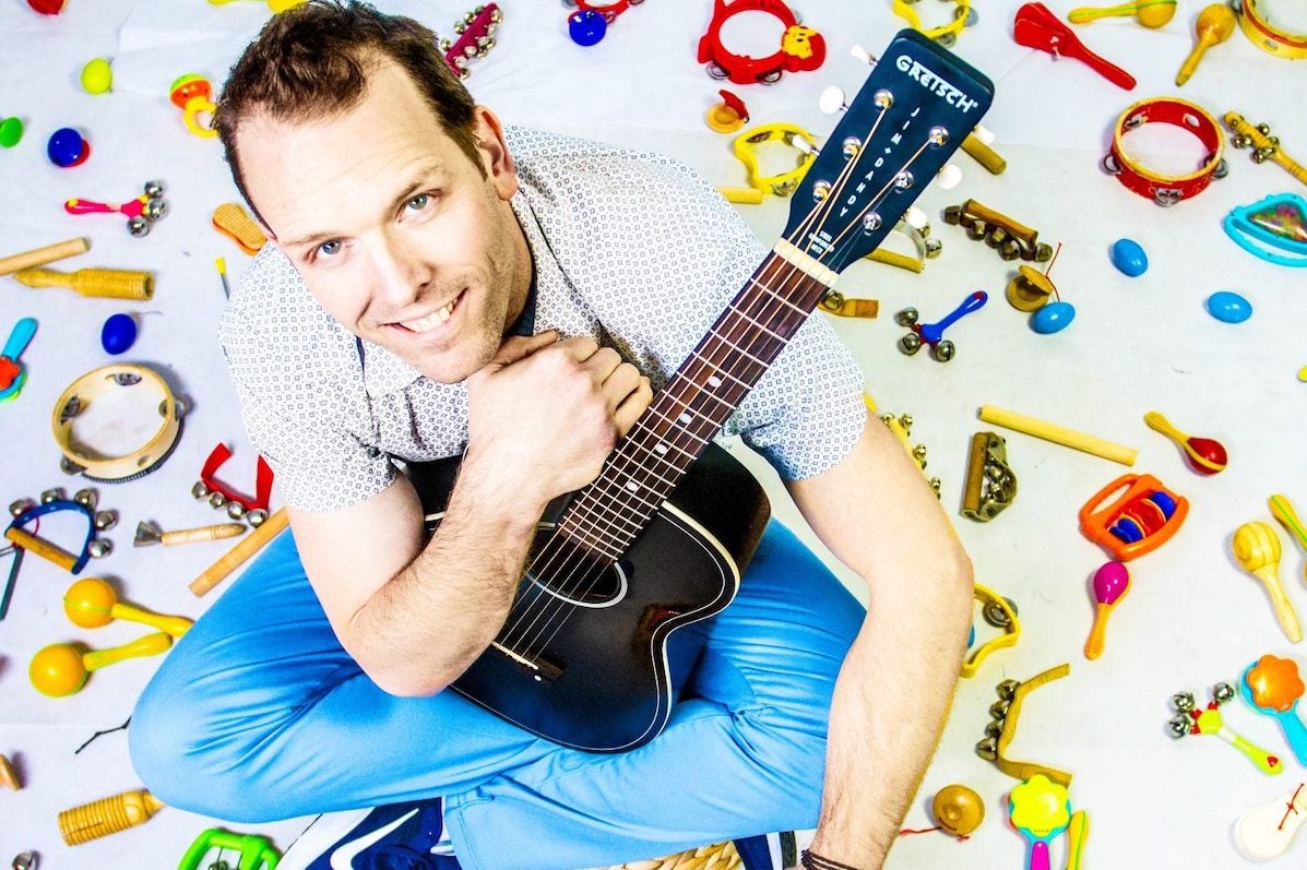 A smiling man holding a black guitar is seated on a white ground surrounded by small, colourful child musical instruments.