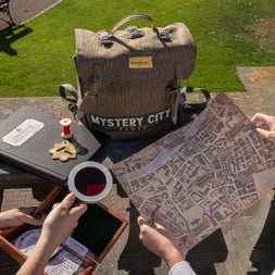 Light brown backpack with a mystery city label on it next to a black notebook with a thread spool and two peoples hands one holding a map and one holding a small box and a magnifying glass