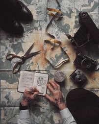 A designer sketching garment ideas in a notepad with his scissors and measuring tape on the table beside a pair of shoes and a camera