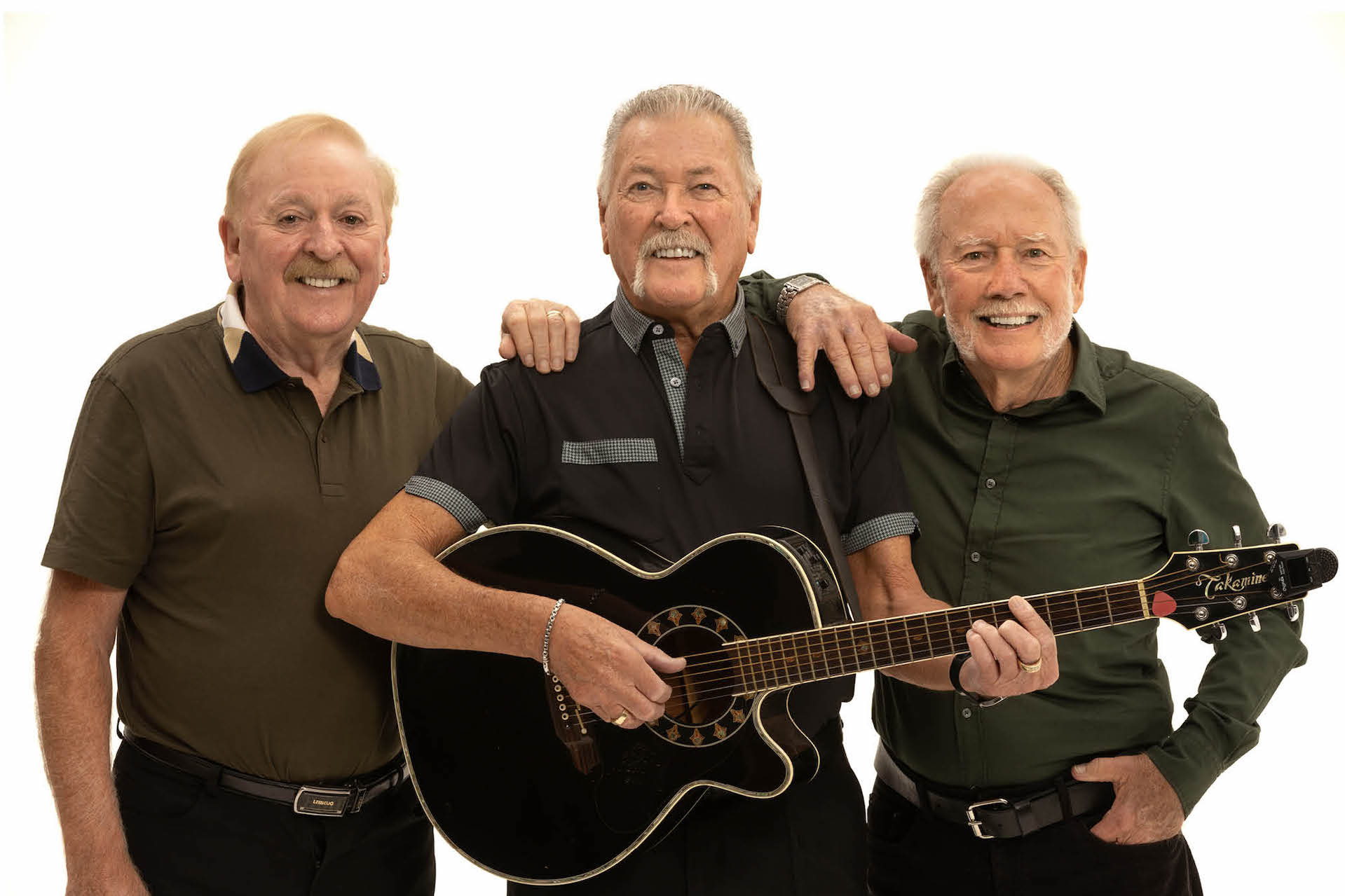 3 older men in a line, the middle one playing a guitar, all smiling, against plain white background.
