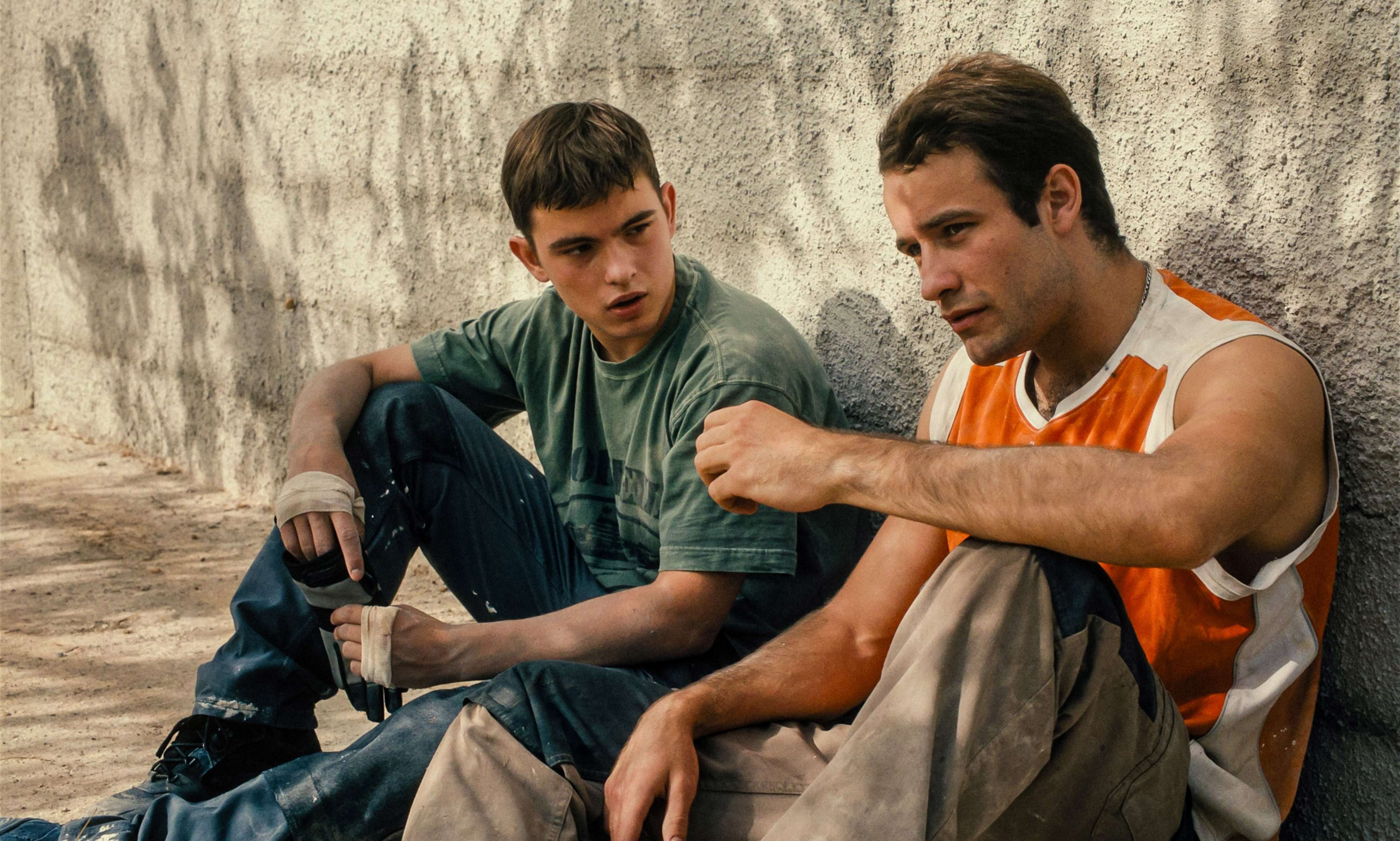 A boy in his late teens and a young man are sitting against a wall.