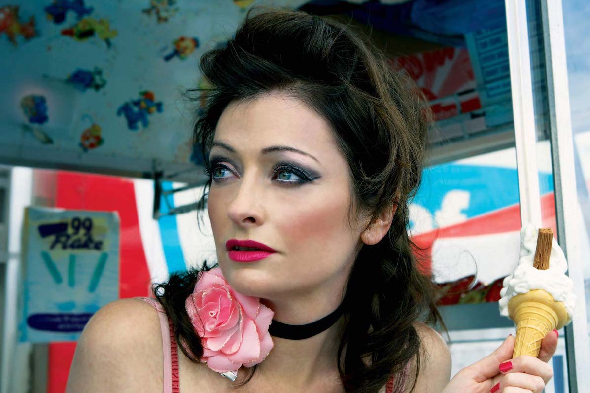 A woman with dark make-up is holding an ice cream looking away to her right.
