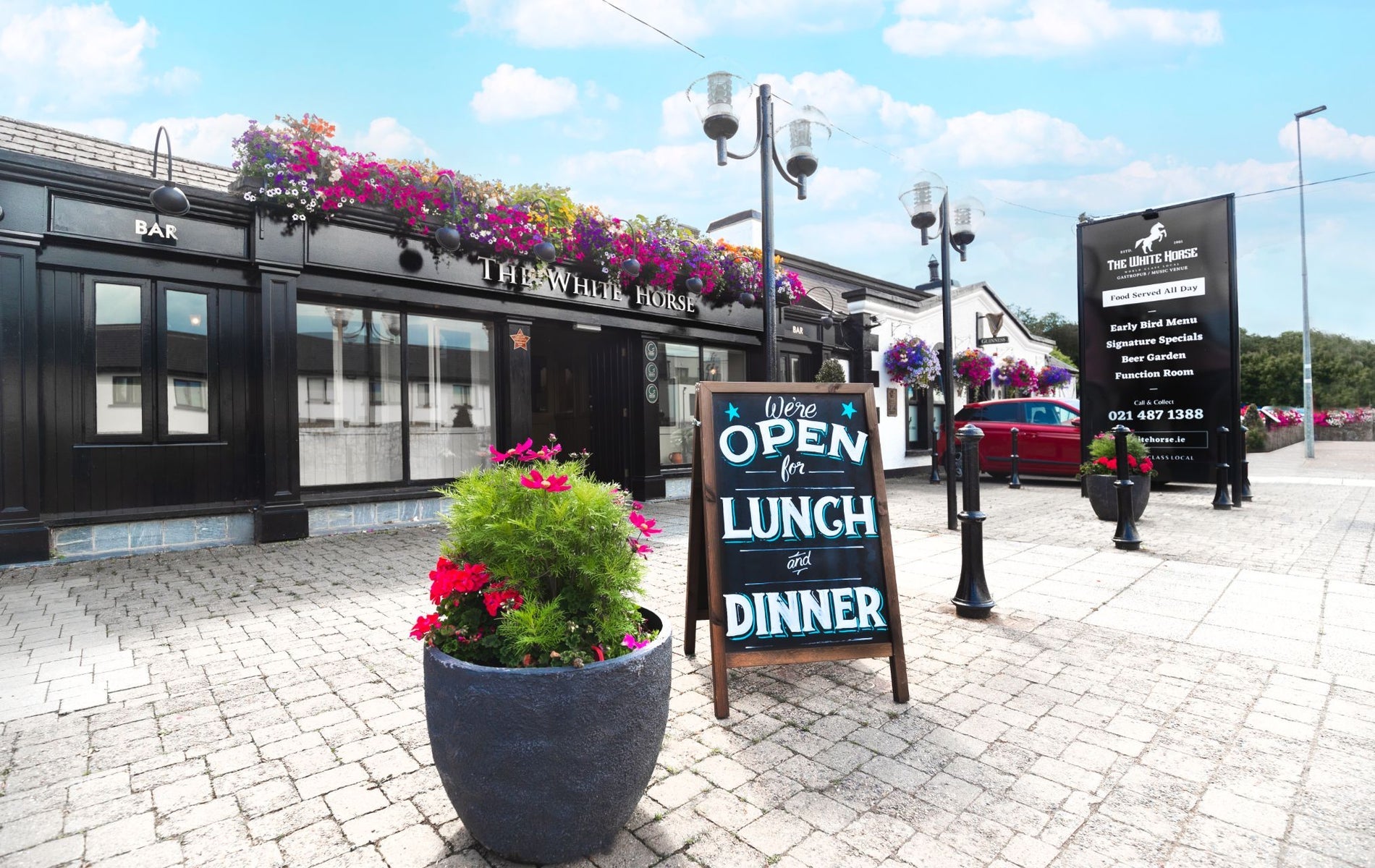 The exterior of a pub with flowers along the front and a lunch sign on the street