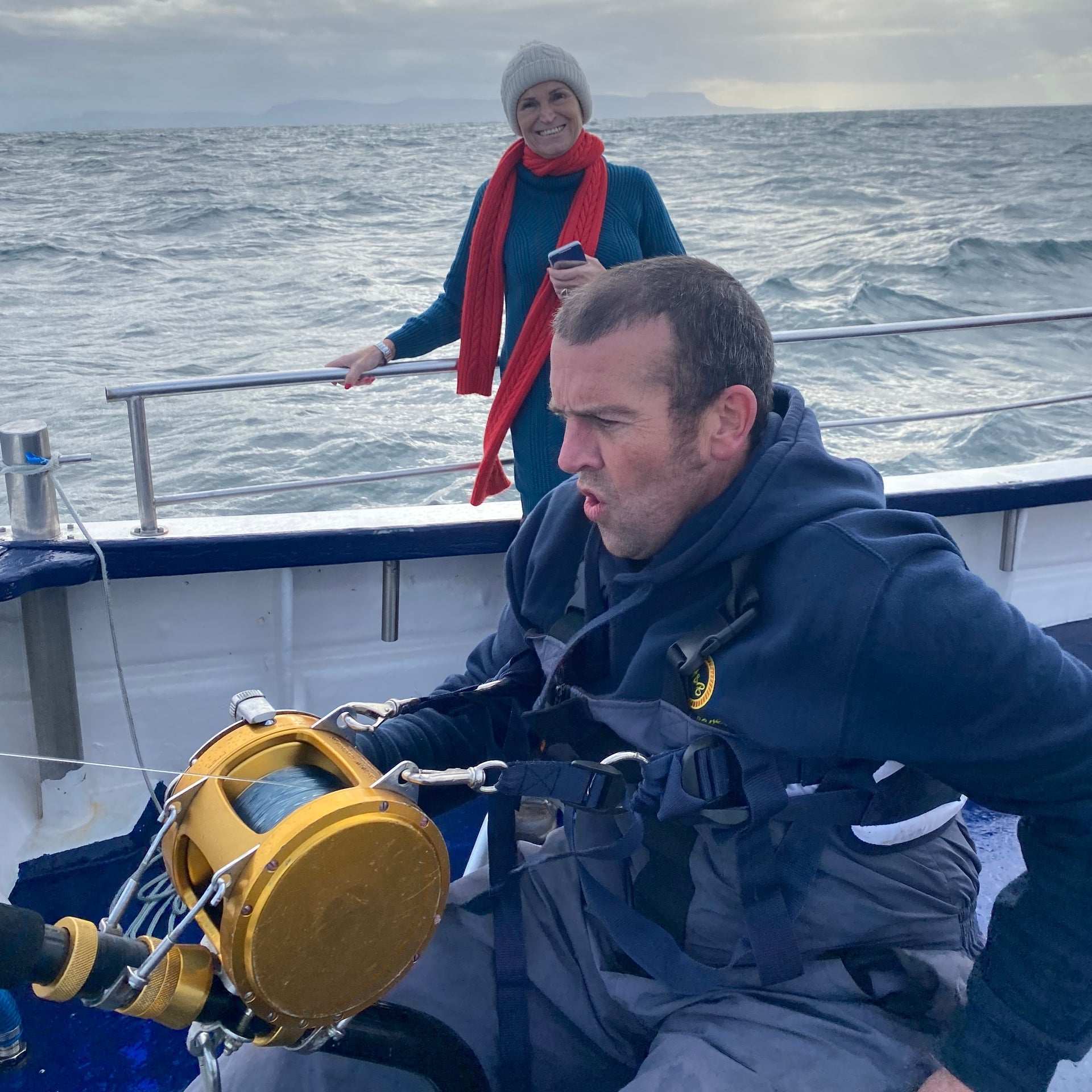 Man on a boat holding a yellow fishing reel with a woman holding a phone in the background