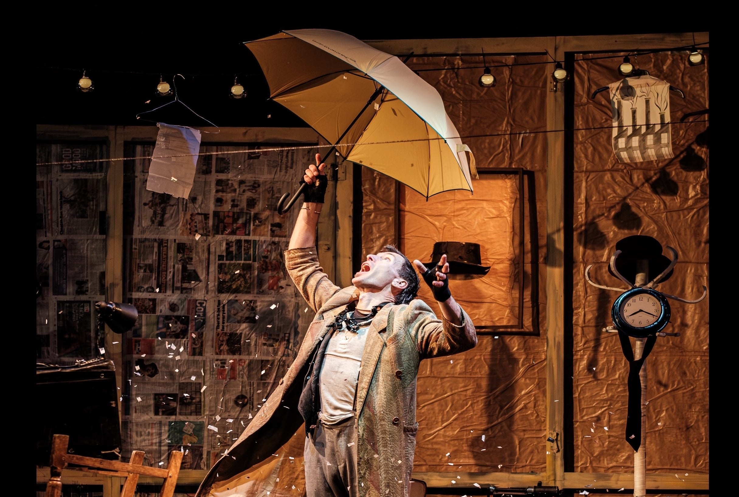 The Collector at An Táin, a man is holding an umbrella over his head on a stage surrounded by busy background stuff.
