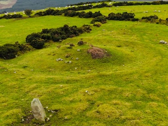 A drone shot of that shows a granite standing stone in the foreground with a burial mound called a barrow behind it in the centre of the image