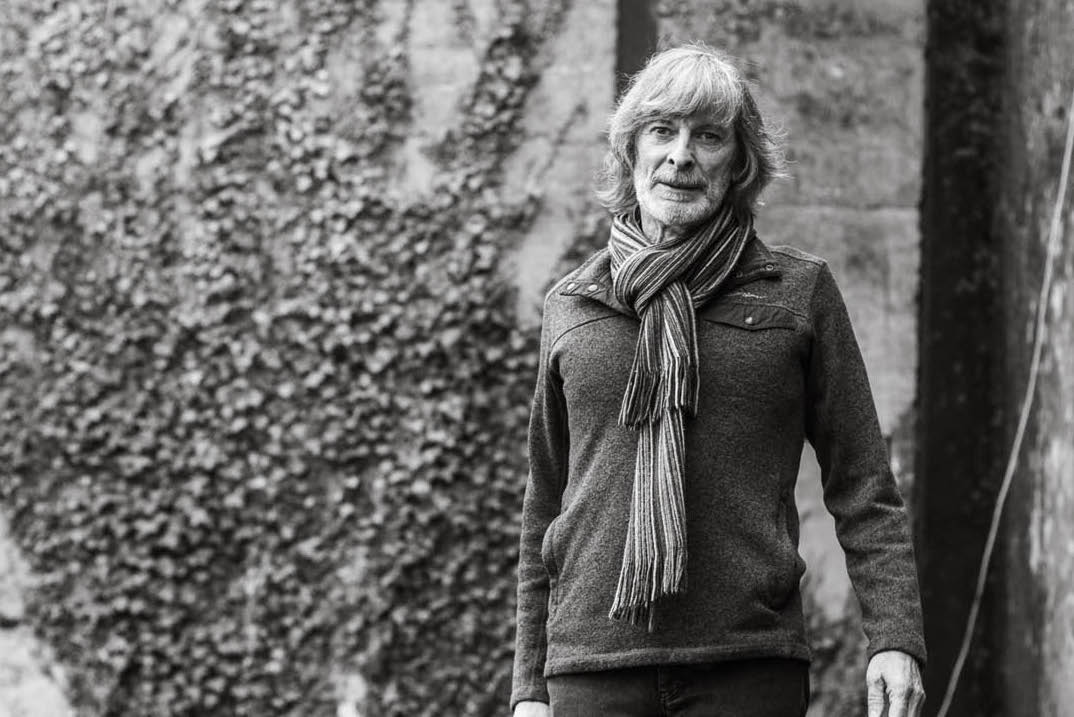 Black and white photo of man wearing a scarf and jumper, standing in front of stone building covered in ivy.
