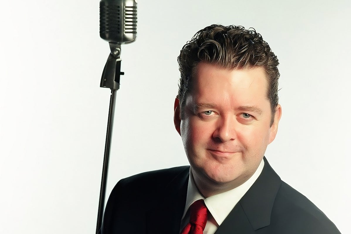 A man in suit and red tie is smiling at the camera, old style large mic is beside him.