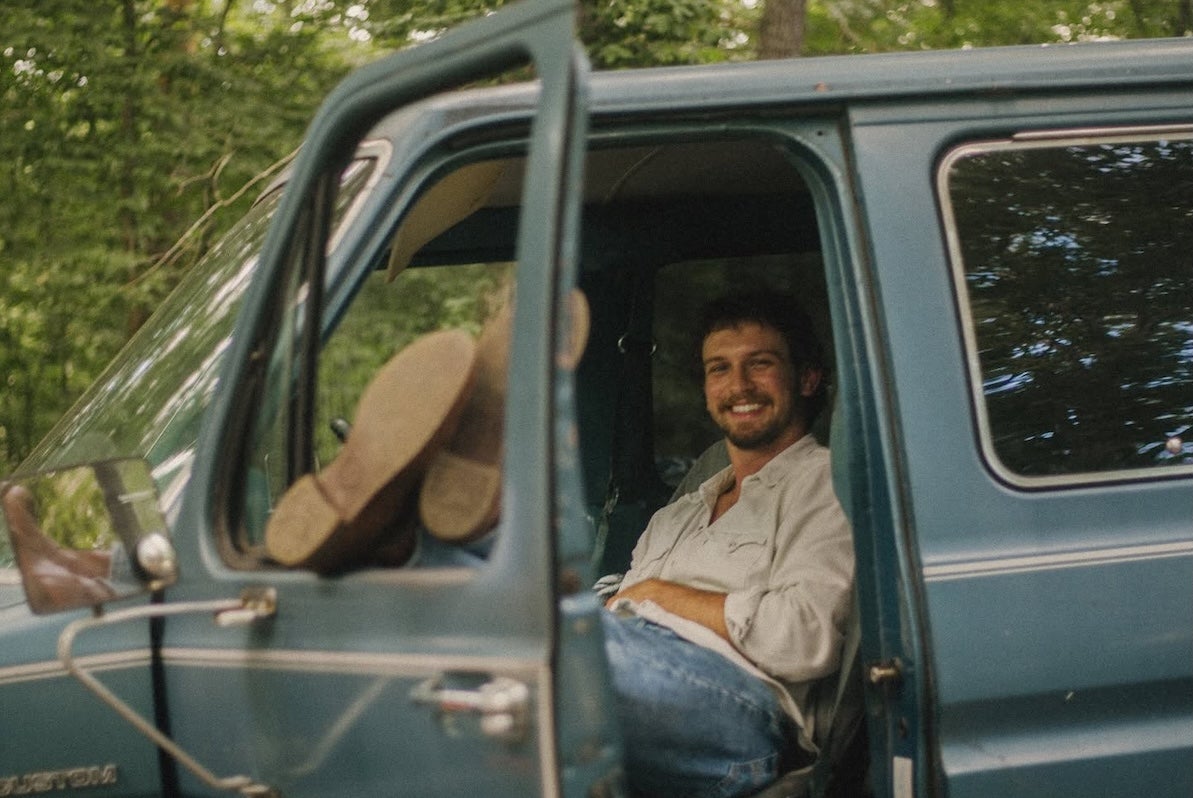A smiling mans seated in an old van with his legs resting up on the open window of an open door.