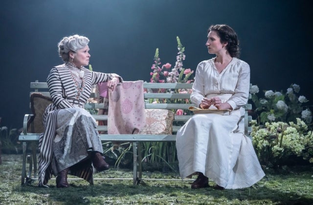 2 women in long dresses are seated on a bench in conversation on a stage