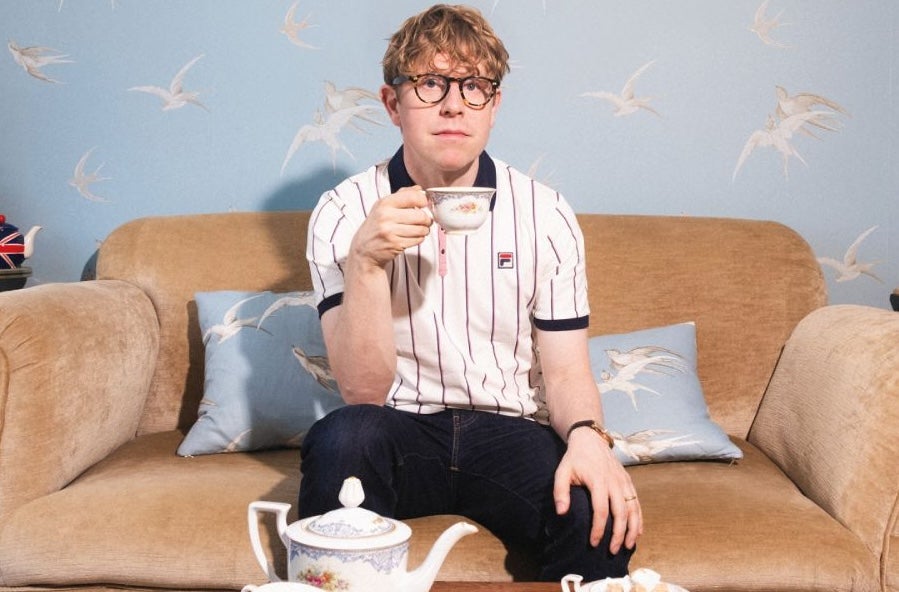 A serious looking man seated on a small sofa holding up a cup of tea with a teapot in front on a low table