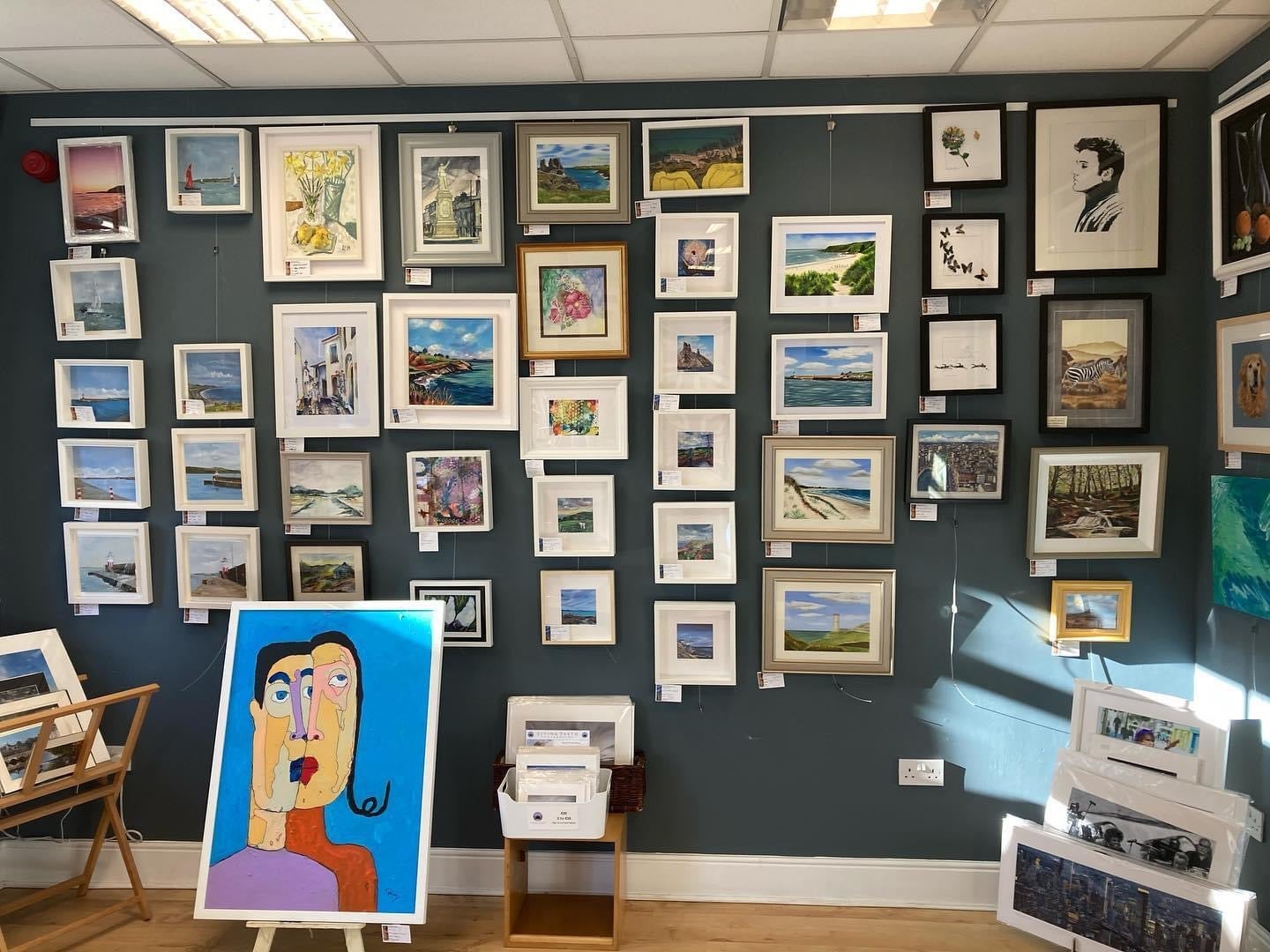 Teal wall with paintings displayed and a white box with prints in front of it