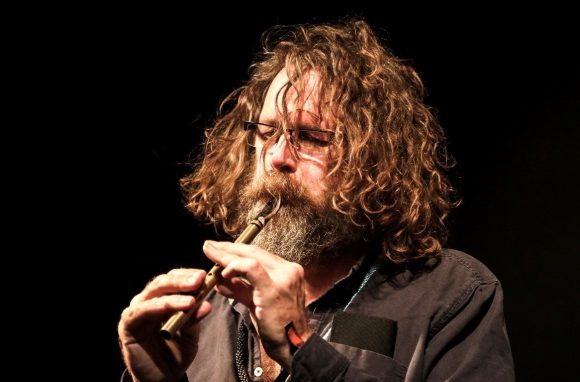 A man with scruffy wavy hair is playing a tin whistle