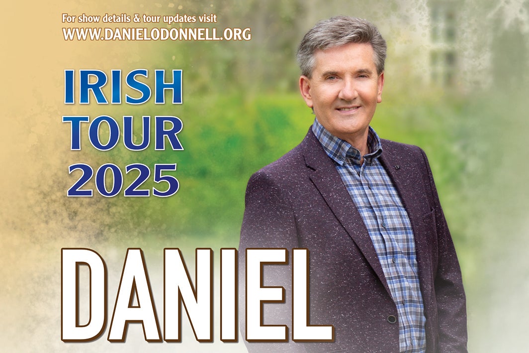 A smiling man in blue checked shirt and dark jacket outdoors with hedge and building in background.