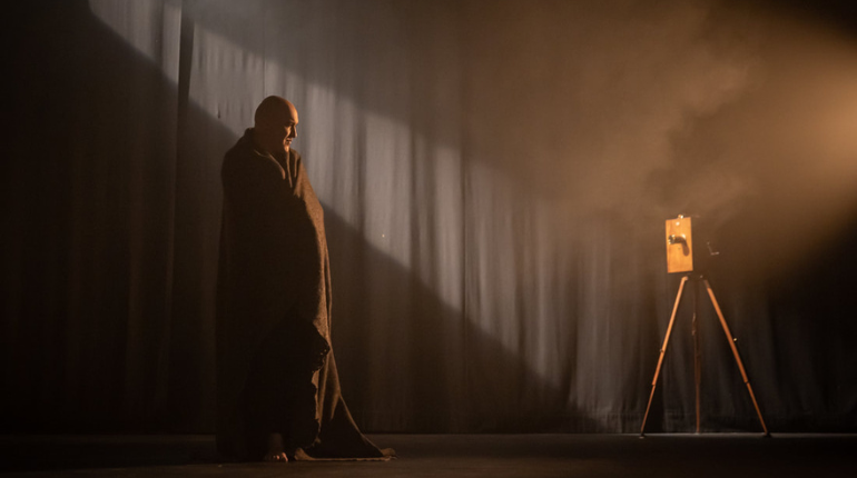 Fishamble presents Silent by Pat Kinevane - a man wrapped in a blanket is looking at old style camera on a tripod