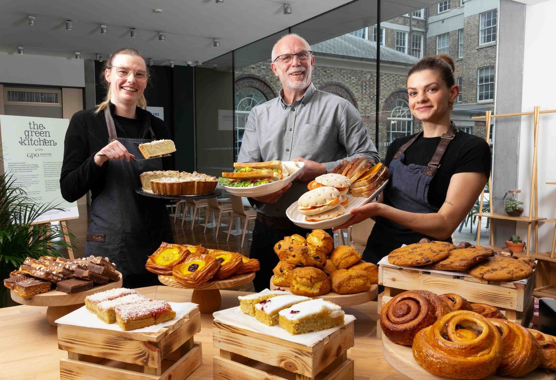 Three people holding food with a display of pastries on a counter in front of them