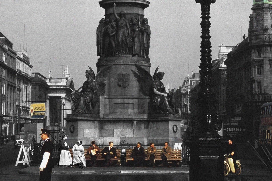 People scattered along 2 benches underneath plinth of large monument in a city in the past.