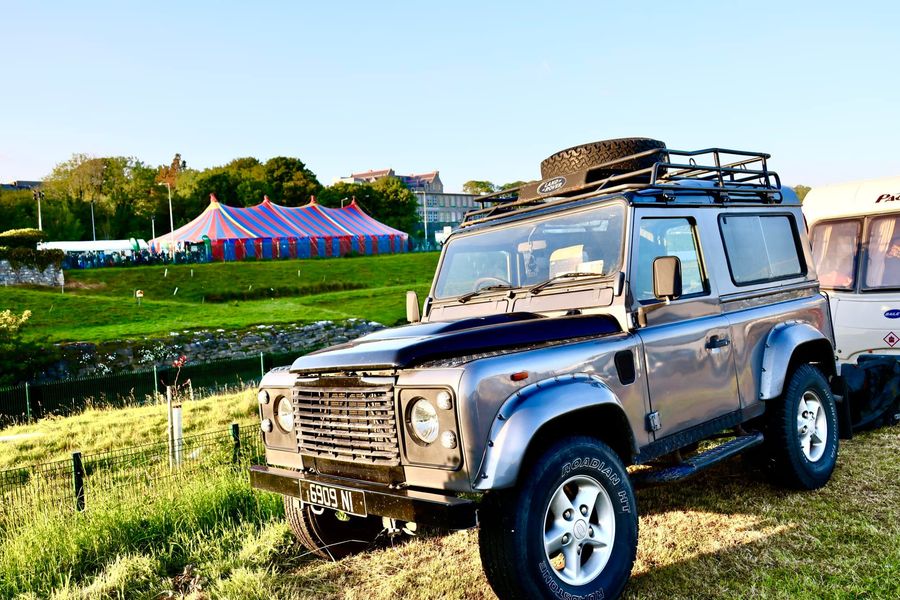 A small shiny jeep car in the foreground, across a green area is a stripy big top tent in the background.