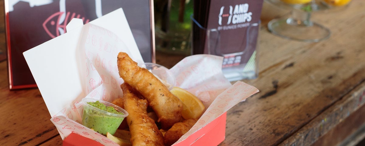 A portion of fish and chips in a box with a lemon slice and dip