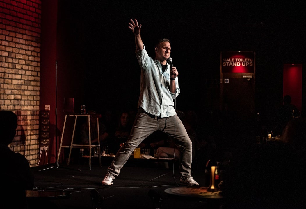 A comedian on stage holding a microphone with his right arm and leg extended