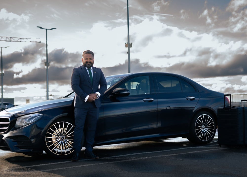 A driver stands beside a dark car in a parking area under a cloudy sky