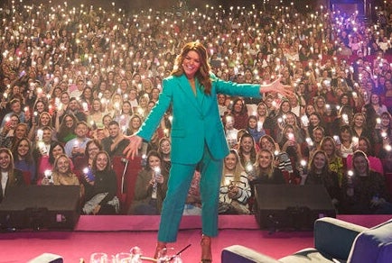 Doireann Garrihy - The Laughs Of Your Life Live, a smiling woman with arms outstretched on a stage with large audience in the background holding lights.