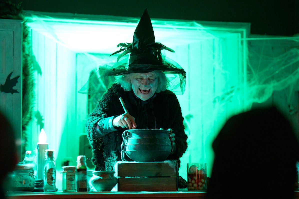 A person wearing black with tall, pointy hat is stirring a small black pot on a steaming block with small bottles, all lit from behind with bright green light.