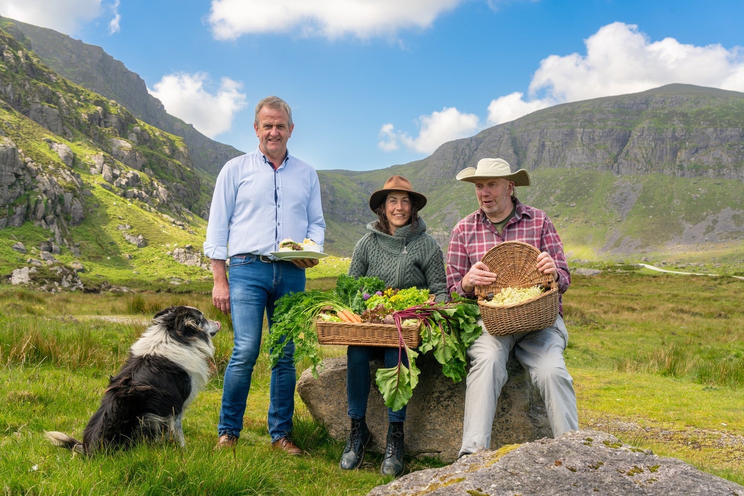 Three people posing with food in baskets and a sheepdog with a mountain in the background
